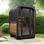 Full-glass front cube sauna under evening sky, exterior lighting aglow