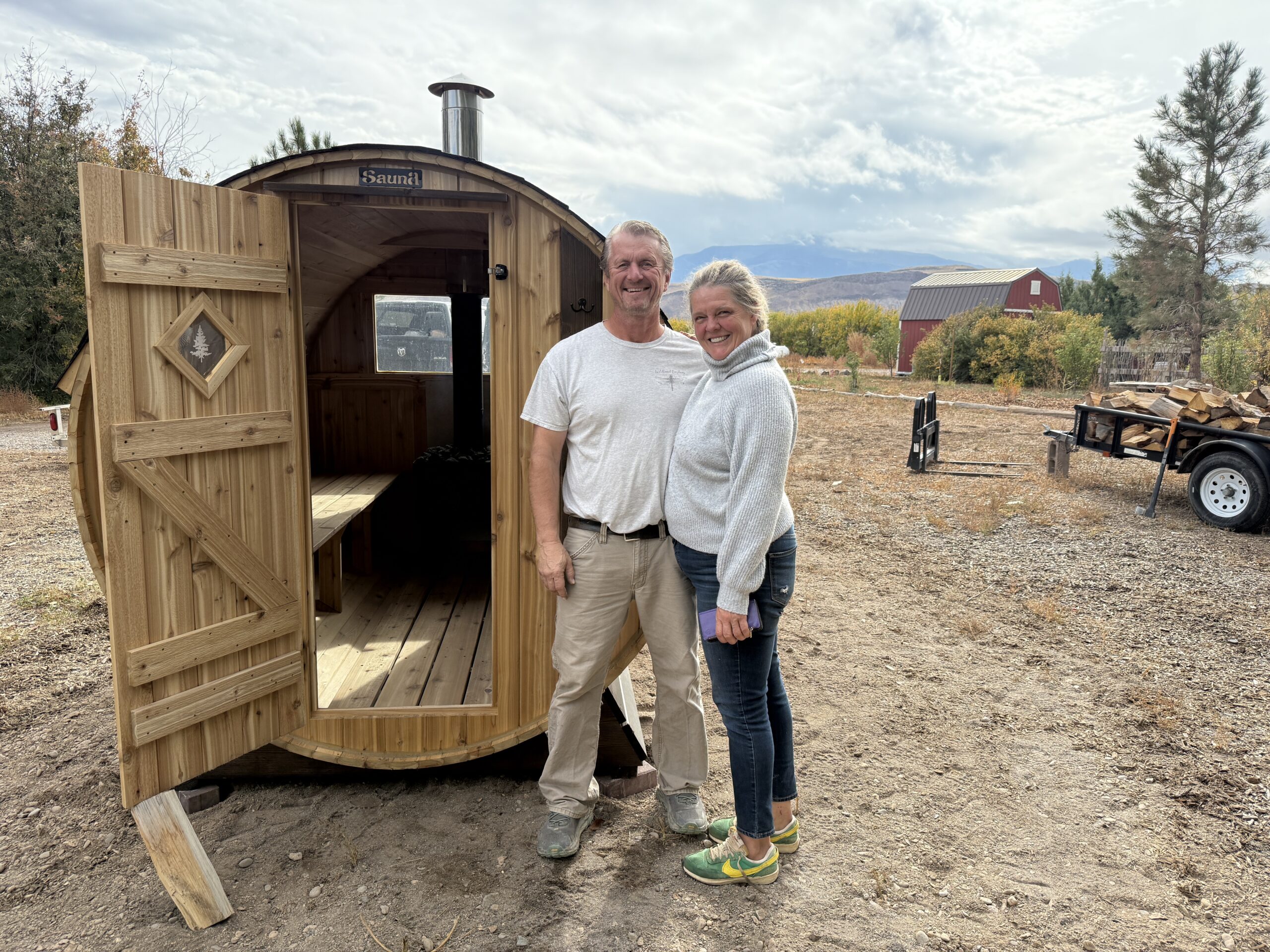 Carrie and her husband standing in front of their Cedar Sense barrel sauna at Wildland Gardens